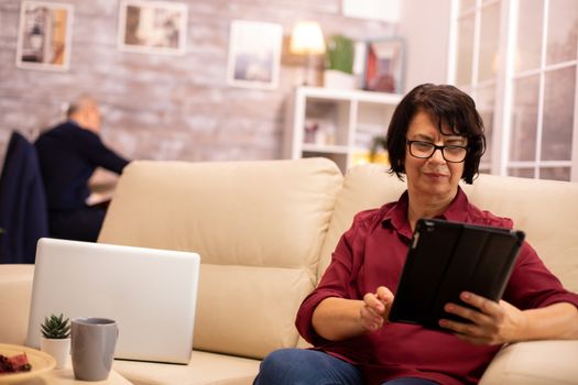 Old elderly woman sitting on the sofa and using a digital tablet PC in cozy living room.