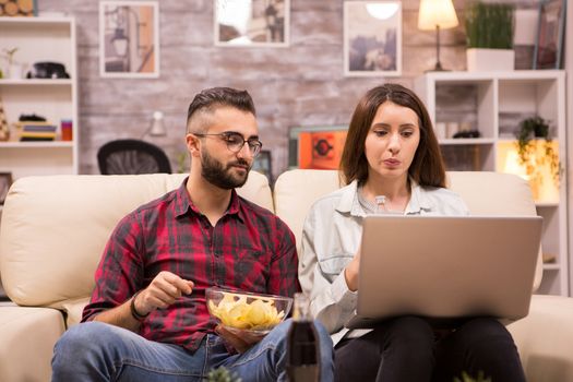 Couple watching a movie sitting comfortable on couch eating chip and drinking soda. Movie on laptop.