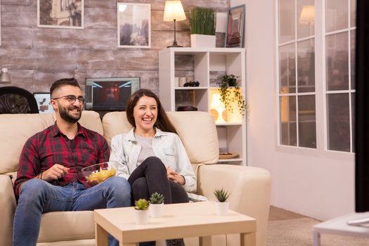 Cheerful young couple sitting on sofa and watching a movie on tv while eating chips and drinking soda.