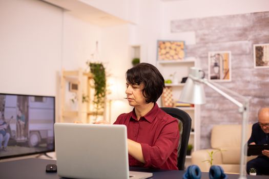 Old woman using a modern computer in her living room while her husband walks in the background