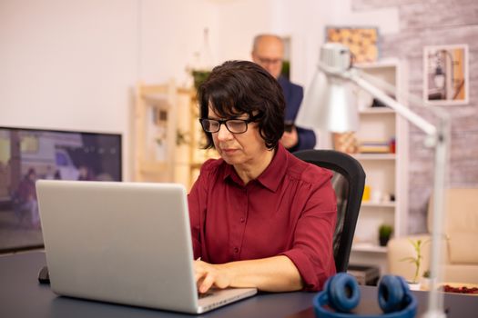 Old woman using a modern computer in her living room while her husband walks in the background