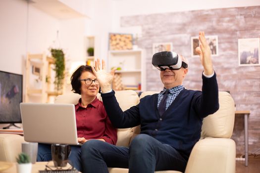 Old elderly retired man using VR virtual reality headset in their cozy apartment.