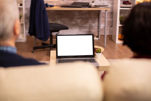 Back view of old couple in their living room looking a laptop with white isolated mock-up.