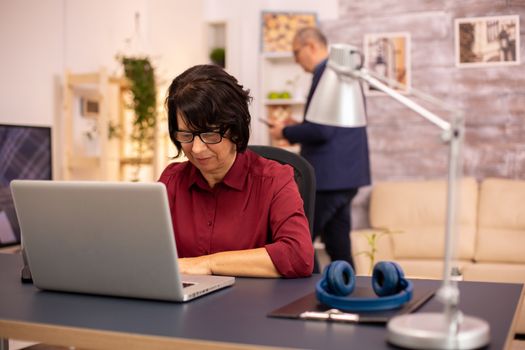 Old woman using a modern computer in her living room while her husband walks in the background