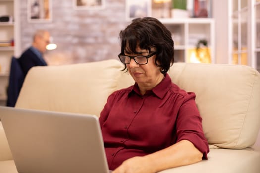 Old elderly woman on her sofa working on a modern laptop in her cozy living room. Her husband is in the background