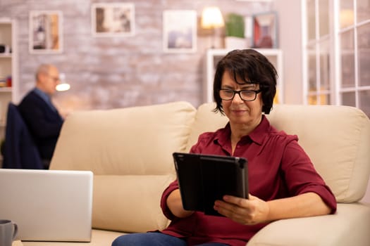 Old elderly woman sitting on the sofa and using a digital tablet PC in cozy living room.