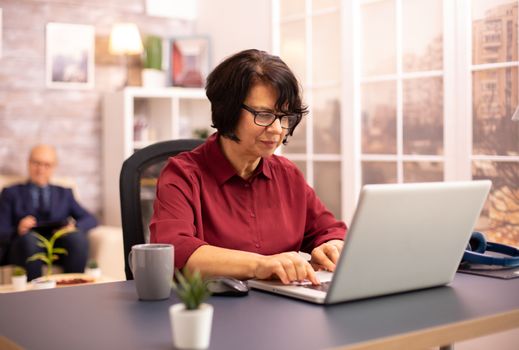 Old woman in her 60s using a modern laptop in her cozy house late in the evening