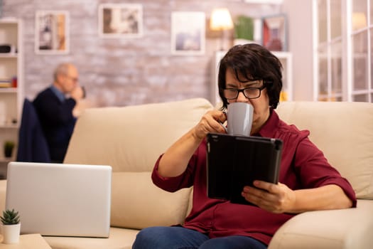 Old elderly woman sitting on the sofa and using a digital tablet PC in cozy living room.