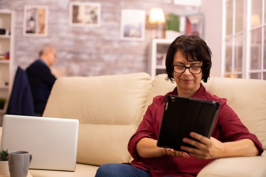 Old elderly woman sitting on the sofa and using a digital tablet PC in cozy living room.