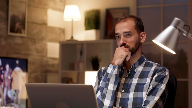 Businessman thinking and holding arms crossed while working on laptop in home office during night hours.