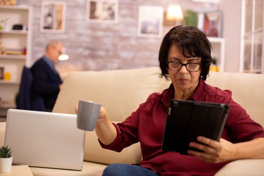 Old elderly woman sitting on the sofa and using a digital tablet PC in cozy living room.