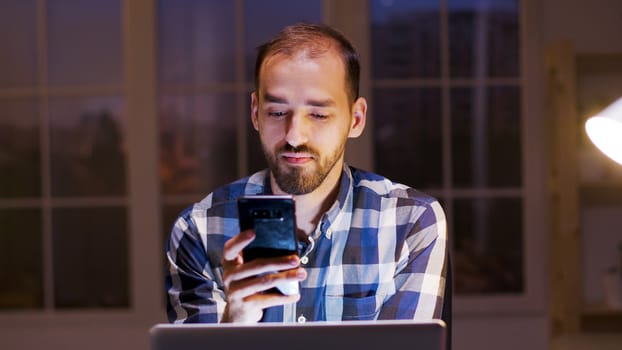 Businessman smiling while looking into his phone during late night work hours in his home office.