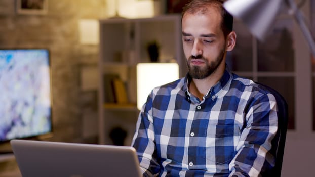 Male freelancer typing on laptop during night hours in his home office.