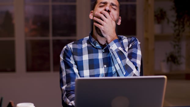 Tired businessman yawning while working on his laptop during night hours in his home office.