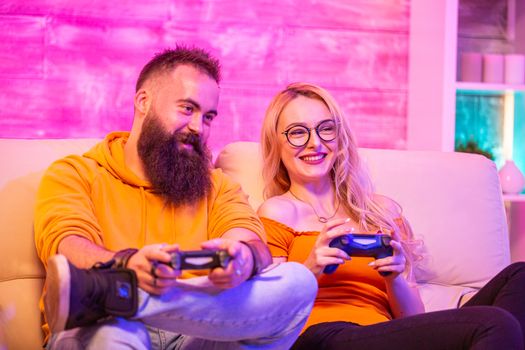 Beautiful blonde girl smiling while playing video games with her boyfriend using wireless controllers on a big screen TV