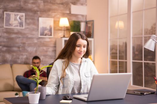 Young woman smiling while working on laptop and holding a cup of coffee. Boyfriend relaxing on sofa in the background.