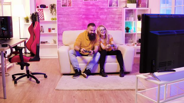 Bearded boyfriend playing online games with his beautiful blonde girlfriend sitting on their couch using wireless controllers.