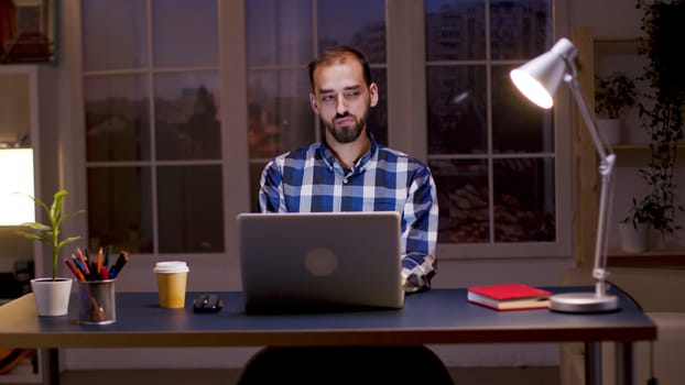 Bearded businessman wearing a shirt and taking a sip of coffee while working from home at night hours.