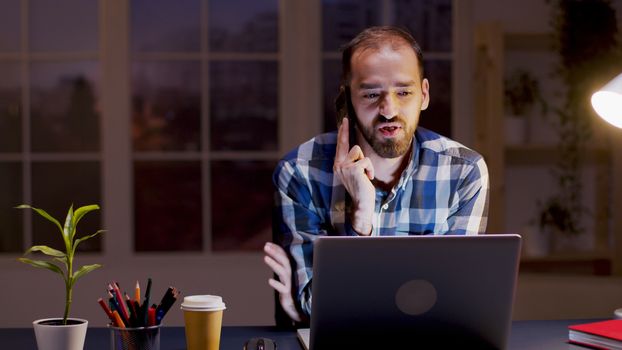 Bearded businessman having a conversation on his phone while working during night hours in his home office.