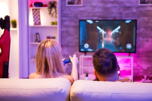 Back view of young couple relaxing playing online shooter games on television using wireless controller.