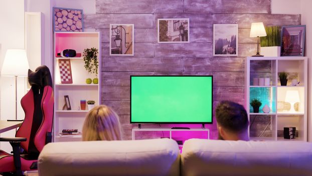 Young couple enjoying playing online games together sitting on sofa. Tv with green screen.