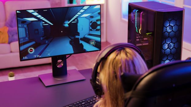 Back view of cheerful young girl playing shooter games sitting on gaming chair. Room with colorful neon lights.