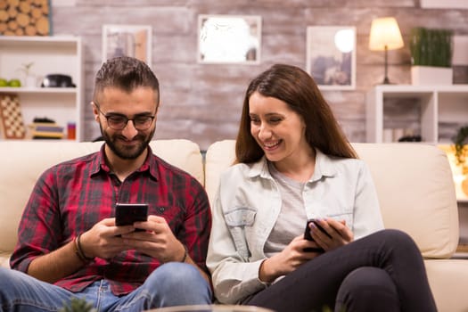 Cheerful young couple laughing together while sitting on sofa. Boyfriend showing something to his girlfriend on phone.
