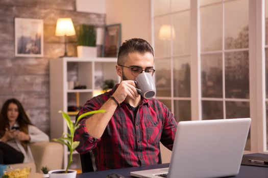 Bearded entrepreneur drinking coffee while working on laptop in living room. Girlfriend relaxing on sofa in the background using phone.