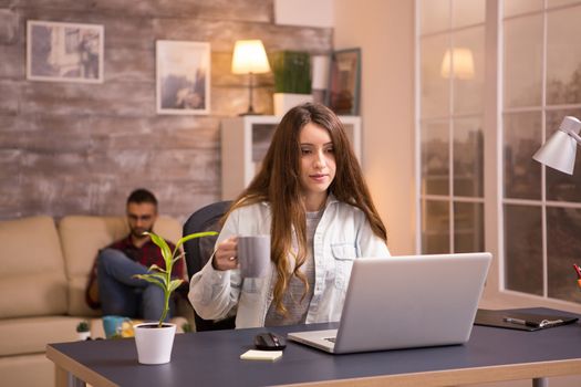 Pretty young girl holding a cup of coffee while working on laptop from home. Man relaxing on sofa and watching something on his phone.