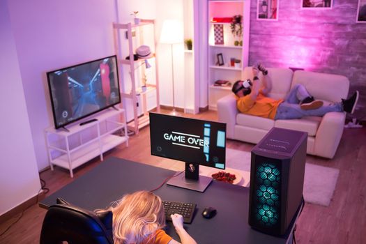 Top view of beautiful blond girl after she lost on computer game. Man with vr goggles on sofa.