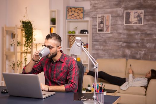 Bearded entrepreneur drinking coffee while working on laptop from home. Girlfriend lying on sofa in the background.
