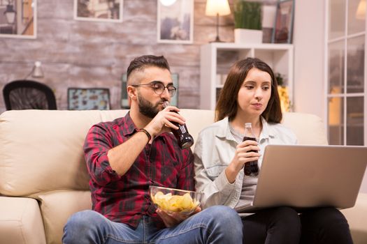 Couple watching a movie sitting comfortable on couch eating chip and drinking soda.