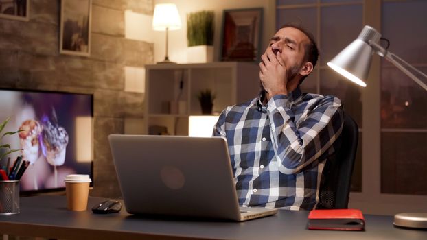 Tired manager yawning while working on laptop in home office during night hours. Overworked businessman.