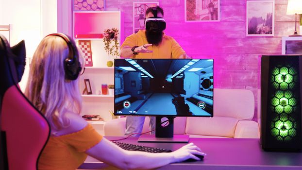 Blonde girl playing online shooter games sitting on gaming chair. Boyfriend wearing virtual reality goggles.