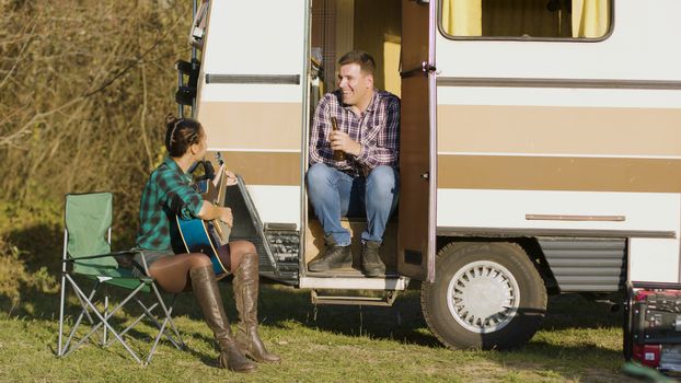 Cheerful woman singing a song on guitar for her boyfriend in front of their retro camper van. Weekend trip.