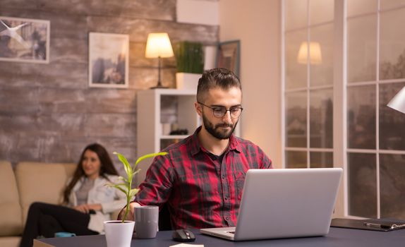 Bearded entrepreneur holding a cup of coffee while working on laptop in living room. Girlfriend relaxing on sofa watching tv in the background.