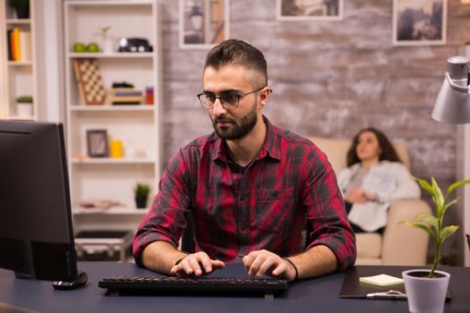 Bearded freelancer typing on computer while working from home. Girlfriend relaxing on sofa in the background.