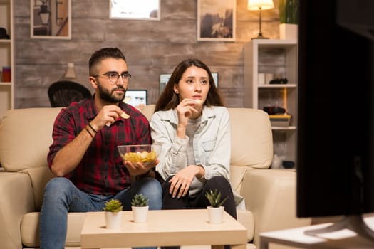 Beautiful young couple looking worried at tv while watching a movie. Couple eating chips sitting on sofa.