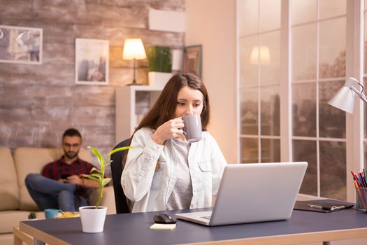 Girlfriend enjoying a cup of coffee while working on laptop from home. Boyfriend relaxing in sofa in the background.