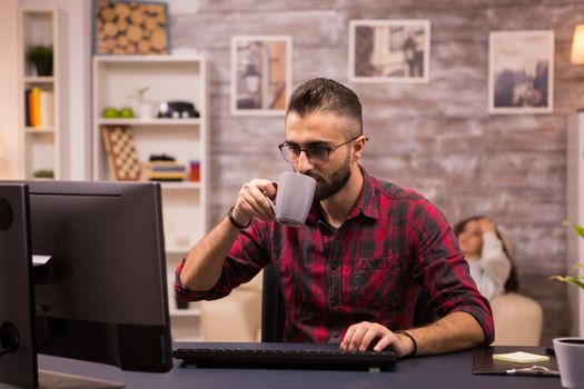 Caucasian entrepreneur taking a sip of coffee while working on computer from home. Girlfriend relaxing on sofa in the background.
