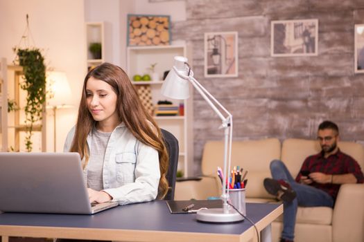 Man relaxing on sofa using his mobile phone while girlfriend is working on her laptop.