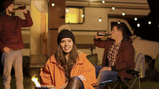 Girl sitting on a camping chair in a cold night of autumn. Friends clinking beer bottles in the background.