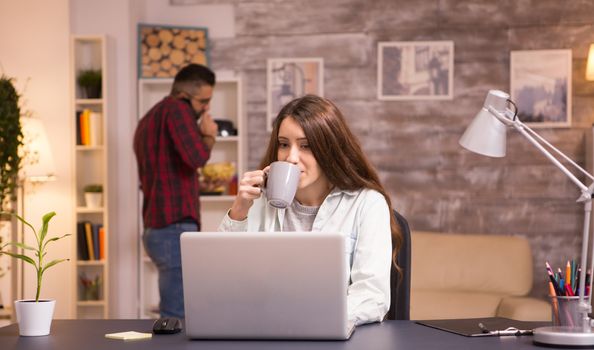 Caucasian female freelancer working on laptop in living room and enjoying a cup of coffee. Boyfriend talking on the phone and eating chips in the background.