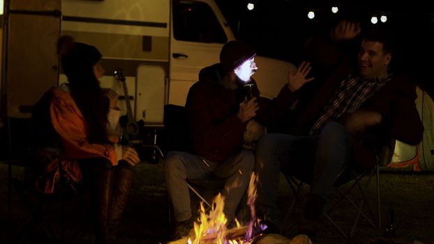 Bearded man telling a scary story to his friends around camp fire in a cold night of autumn. Retro camper van. Light bulbs