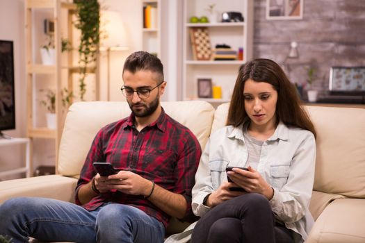 Caucasian couple with serious expressions using their phones while sitting on couch.