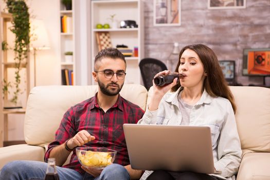 Girlfriend drinking soda while boyfriend's eating chips sitting on sofa and watching a movie.
