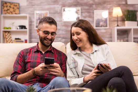 Caucasian boyfriend laughing with his girlfriend while showing her something on his phone. Couple sitting on couch