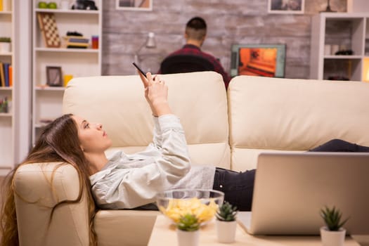 Beautiful young woman laying on sofa end browsing on phone. Boyfriend in the background.