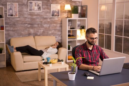 Male entrepreneur working on laptop in living room with girlfriend relaxing on sofa in the background. Chips on table.