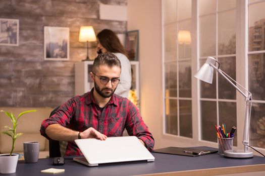 Bearded entrepreneur opening laptop to start working from home. Girlfriend in the background.